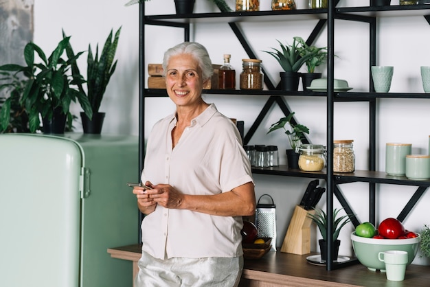 Gros plan d'une femme senior souriante à l'aide de commandes mobiles dans la cuisine