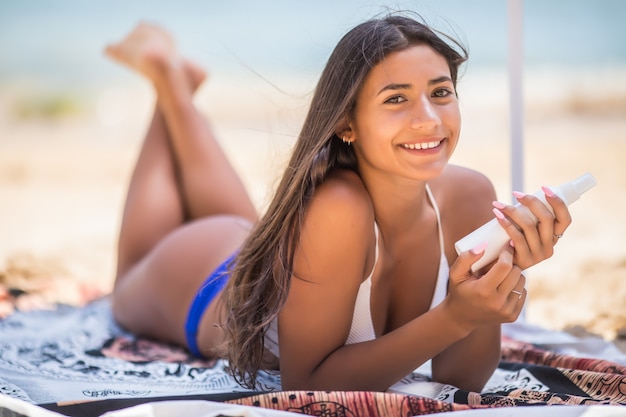 Gros plan d'une femme sexy en vaporisant un écran solaire, une lotion ou un écran solaire sur le corps pour éviter les rayons UV du soleil avant le bain de soleil en été ou des vacances avec la mer et le bluesky