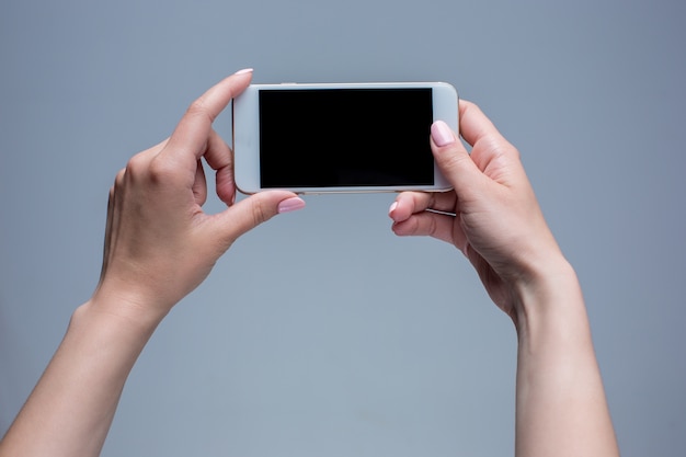 Gros plan d'une femme tapant sur téléphone mobile sur fond gris. Mains féminines tenant un smartphone moderne et pointant avec le doigt.