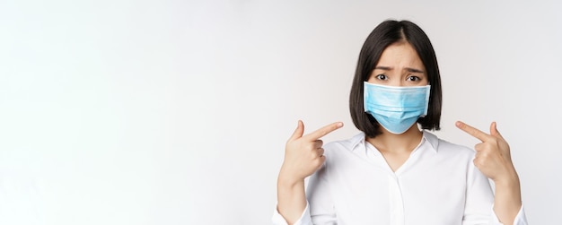 Gros plan d'une fille asiatique triste dans un masque médical pointant vers sa tête et regardant bouleversée debout sur fond blanc