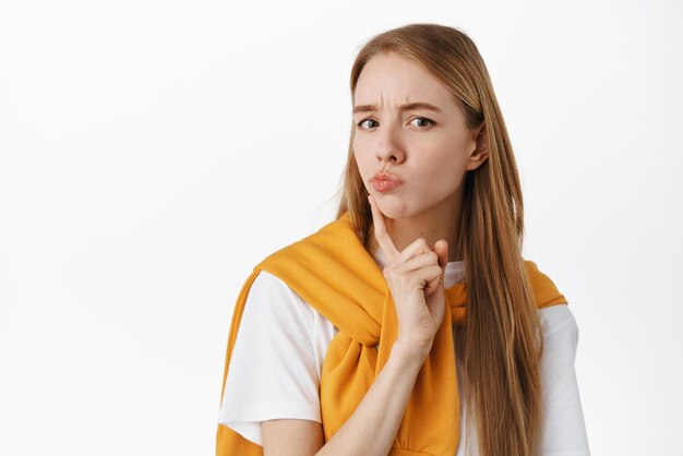 Gros plan d'une fille blonde réfléchie fronçant les sourcils regardant la caméra intriguée en pensant à quelque chose de prendre une décision debout sur fond blanc