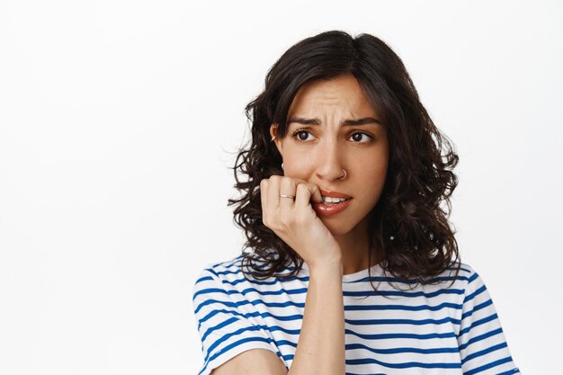 Gros plan sur une fille brune anxieuse, se rongeant les ongles et détournant les yeux avec un visage nerveux et alarmé, ayant peur des conséquences, ayant peur, debout sur du blanc