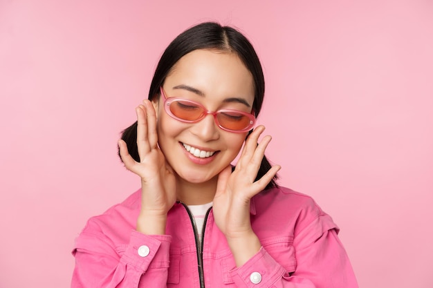 Gros plan d'une fille coréenne élégante à lunettes de soleil souriant heureux posant sur fond rose Les gens font face au concept