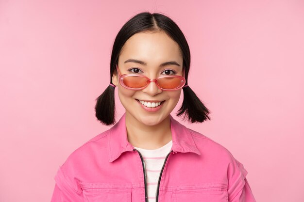 Gros plan d'une fille coréenne élégante à lunettes de soleil souriant heureux posant sur fond rose Les gens font face au concept
