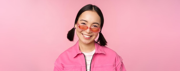 Gros plan d'une fille coréenne élégante à lunettes de soleil souriant heureux posant sur fond rose Les gens font face au concept