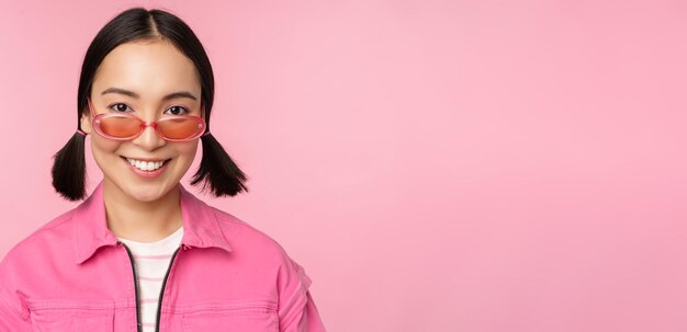 Gros plan d'une fille coréenne élégante à lunettes de soleil souriant heureux posant sur fond rose Les gens font face au concept