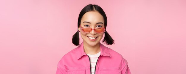 Gros plan d'une fille coréenne élégante à lunettes de soleil souriant heureux posant sur fond rose Les gens font face au concept