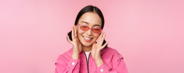 Gros plan d'une fille coréenne élégante à lunettes de soleil souriant heureux posant sur fond rose Les gens font face au concept