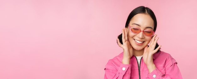 Gros plan d'une fille coréenne élégante à lunettes de soleil souriant heureux posant sur fond rose Les gens font face au concept