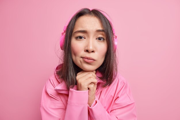 Gros plan d'une fille millénaire triste et déçue qui garde les mains sous le menton avec une expression mélancolique écoute des paroles de chansons via des écouteurs vêtus d'une veste isolée sur un mur rose