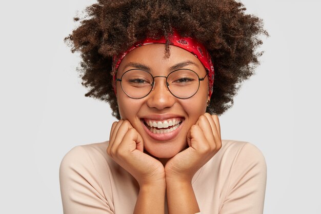 Gros plan d'une fille à la peau sombre touche les joues, se sent heureuse, apprécie une musique agréable et une bonne journée, porte de grandes lunettes rondes, des modèles sur un mur blanc. Femme féminine sensuelle sourit largement