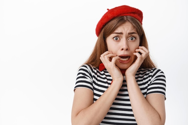 Gros plan sur une fille qui a peur, tremble de peur, se ronge les ongles et a l'air anxieux devant la caméra, debout dans un béret rouge français et un t-shirt rayé, fond blanc