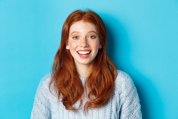 Gros plan d'une fille rousse heureuse en pull, regardant la caméra avec un sourire plein d'espoir, debout sur fond bleu.