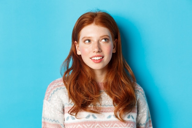 Gros plan d'une fille rousse rêveuse imaginant quelque chose, regardant le coin supérieur droit et souriant, debout dans un pull d'hiver sur fond bleu