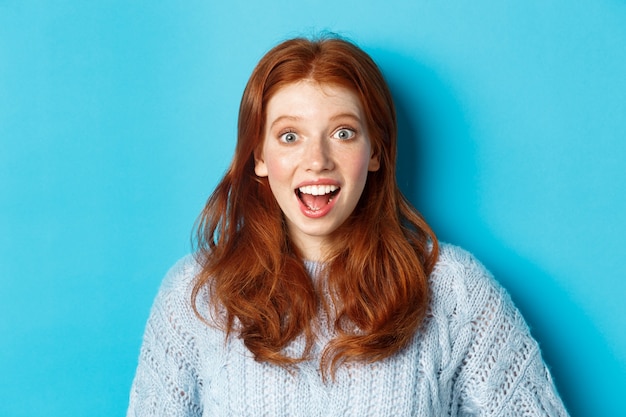 Gros plan d'une fille rousse surprise et excitée qui entend des nouvelles incroyables, regardant la caméra, regardant l'offre promotionnelle, debout sur fond bleu.