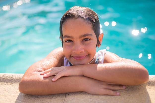 Gros Plan D'une Fille Souriante Sur Fond D'eau Bleu Pétillant. Fille Caucasienne En Maillot De Bain Debout Dans La Piscine Penchant Son Menton Sur Les Mains Mouillées Et Regardant La Caméra. Repos Actif Et Concept D'enfance Insouciante