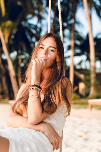 Gros plan d'une fille tendre assise sur une balançoire vêtue d'une robe blanche. Elle a de longs cheveux noirs. Elle a des bracelets sur son bras. La balançoire est sur la plage avec des palmiers verts