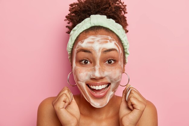 Gros plan de l'heureuse femme afro-américaine bouclée impressionnée lève les poings serrés, prend plaisir à des traitements hygiéniques, porte un bandeau sur la tête, se lave le visage avec du savon à bulles, sourit largement