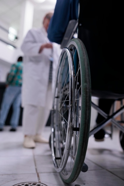 Gros plan sur un homme âgé vivant avec un handicap utilisant un fauteuil roulant à la réception d'une clinique privée discutant avec un médecin d'un rendez-vous. Mise au point sélective sur la roue du fauteuil roulant dans le hall de l'hôpital très fréquenté.