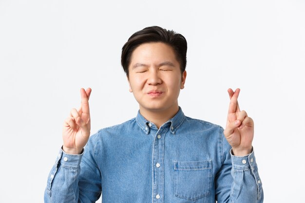 Gros plan d'un homme asiatique inquiet plein d'espoir fermer les yeux et croiser les doigts pour la bonne chance, faire des vœux, prier en attendant les résultats, anticiper les nouvelles, debout mur blanc