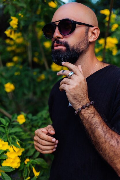 Gros plan d'un homme barbu bronzé brutal à lunettes de soleil ayant tatouage de palmier sur le doigt se dresse entouré de fleurs jaunes dans le parc