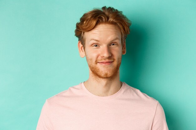 Gros plan d'un homme barbu rousse à la recherche de plaisir, d'un signe de tête d'approbation et souriant, debout en t-shirt rose sur fond turquoise.