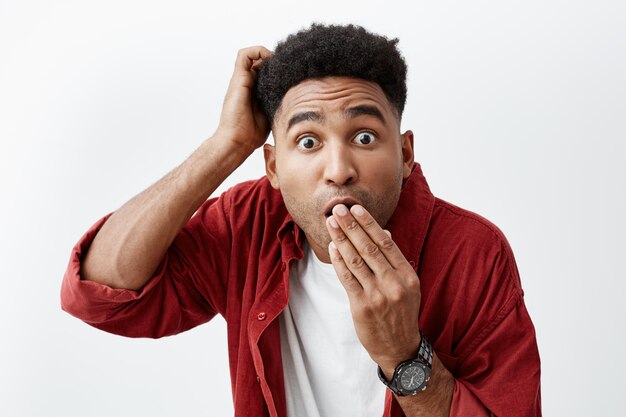 Gros plan d'un homme mature séduisant à la peau noire avec une coiffure afro dans une bouche de vêtements à la mode décontractée avec la main, regardant à huis clos avec une expression choquée après avoir entendu des ragots sur un ami.