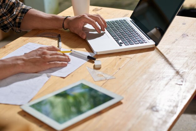 Gros plan d'un homme recadré esquissant un projet sur la feuille de papier et à l'aide de l'ordinateur portable