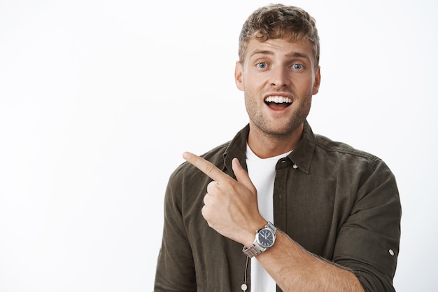 Gros plan d'un homme séduisant et charismatique aux cheveux blonds, aux poils et aux yeux bleus posant une question avec un sourire curieux et excité pointant vers le coin supérieur gauche contre un mur gris