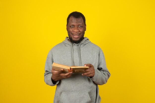 Gros plan sur un homme souriant afro-américain, lisant un livre à la main, se dresse sur un mur jaune.