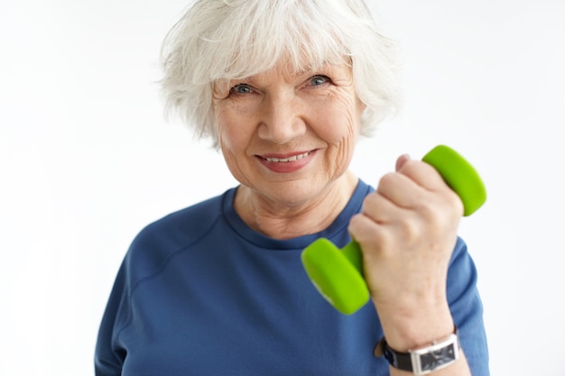 Gros plan image de femme mature sportive énergique avec les cheveux gris et les rides exercice à l'intérieur, faire des boucles de biceps, tenant un haltère vert et souriant joyeusement. Sports, âge et fitness