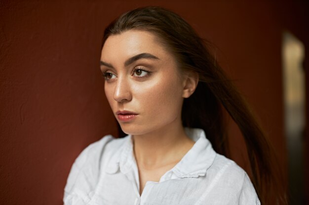 Gros plan image de jeune femme élégante confiante en chemise blanche marchant sur fond de mur brun, vent soufflant ses longs cheveux lâches autour.