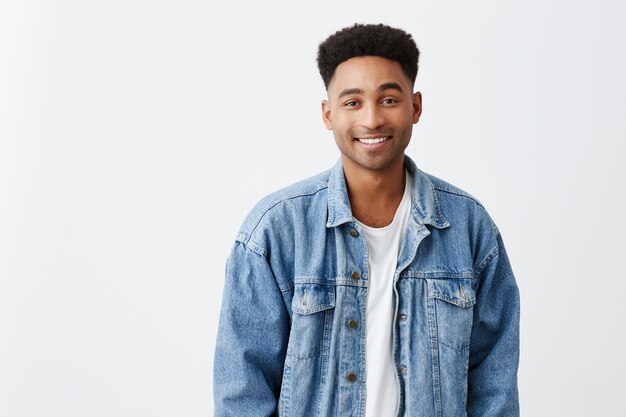 Gros plan isolé sur blanc portrait de jeune belle étudiante de sexe masculin à la peau foncée et gaie avec une coiffure afro en t-shirt blanc et veste en jean souriant brillamment à huis clos. Espace copie