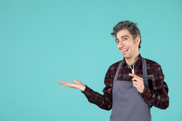 Gros plan d'un jeune coiffeur souriant portant un tablier gris et tenant un peigne et des ciseaux pointant quelque chose sur le côté droit sur fond bleu