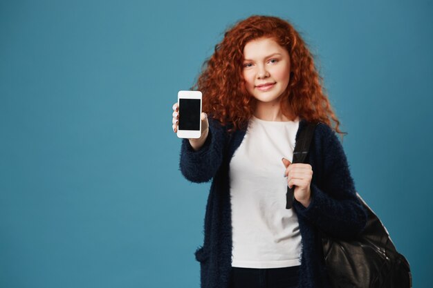 Gros plan d'une jeune étudiante au gingembre avec des cheveux ondulés et des taches de rousseur portant un t-shirt blanc et un cardigan noir montrant un smartphone.