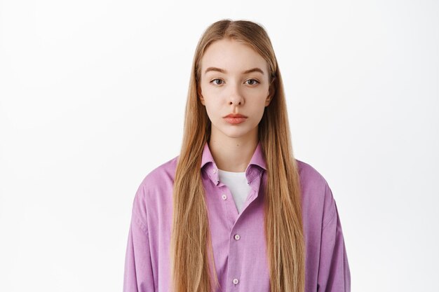 Gros plan sur une jeune femme aux cheveux longs blonds naturels, ayant l'air décontractée sans émotion, pokerface, regardant la caméra, debout sur fond blanc