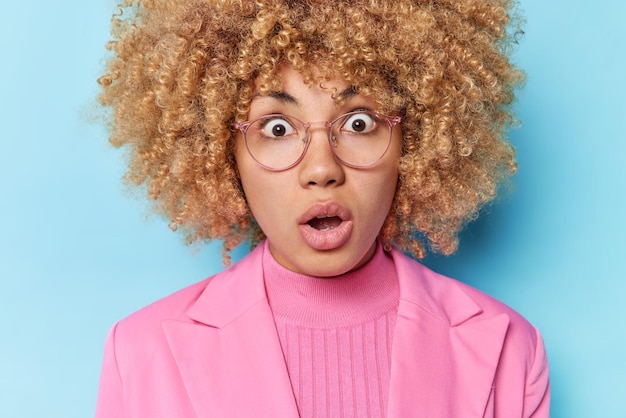 Gros plan d'une jeune femme choquée aux cheveux bouclés garde la bouche ouverte ne peut pas croire en des nouvelles incroyables découvre une pertinence incroyable porte une veste rose isolée sur fond bleu Concept Omg