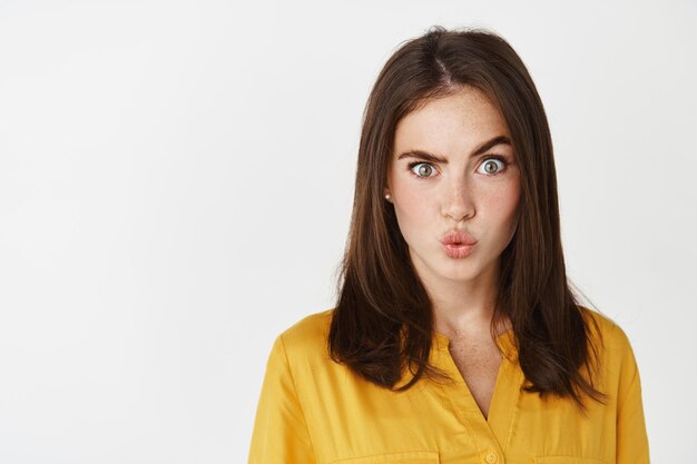 Gros plan d'une jeune femme confuse et intriguée levant un sourcil, plissant les lèvres et regardant l'avant perplexe, debout sur un mur blanc.