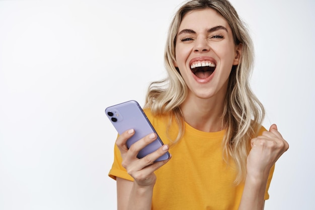 Gros plan sur une jeune femme gagnante riant et souriant à l'aide d'un smartphone et célébrant la victoire gagnante sur fond blanc de téléphone portable