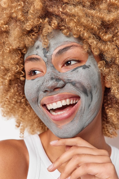 Gros plan d'une jeune femme heureuse et positive avec des cheveux bouclés blonds touche la mâchoire applique doucement un masque d'argile d'argile sur le visage pour réduire les ridules et les pores regarde avec joie porte un t-shirt décontracté