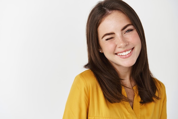 Gros plan d'une jeune femme mignonne et rêveuse souriante, faisant un clin d'œil à l'avant pour encourager le client, debout sur un mur blanc