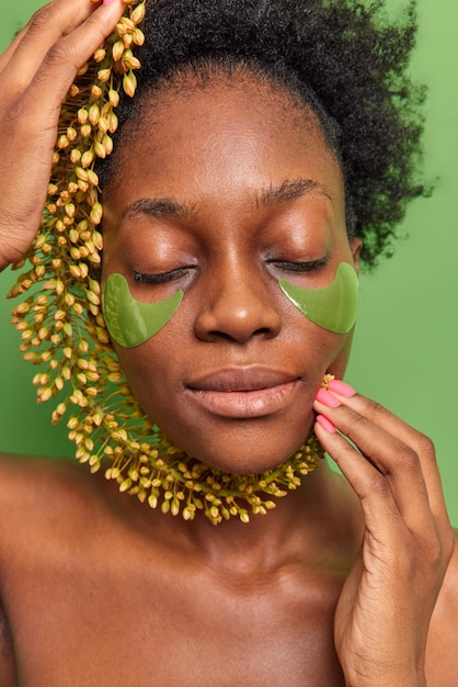 Gros plan d'une jeune femme à la peau foncée qui garde les yeux fermés tient une fleur sauvage sur le visage a une peau saine et lisse avec un corps nu isolé sur fond vert. Concept de femmes et de beauté.