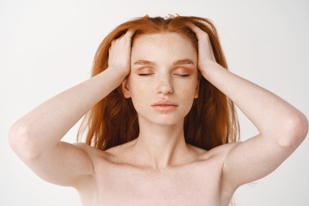 Gros plan d'une jeune femme rousse détendue à la peau pâle et aux taches de rousseur, massant les cheveux roux naturels avec les yeux fermés, debout nue sans maquillage sur un mur blanc
