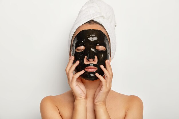 Gros plan d'une jeune femme sérieuse porte un masque facial noir, a les yeux fermés, touche doucement le visage, porte une serviette enveloppée autour de la tête