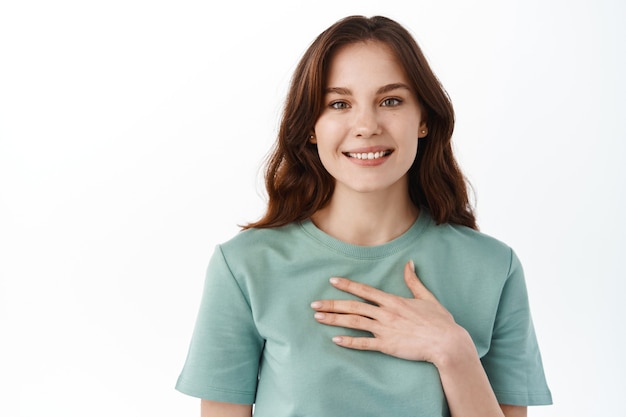 Gros plan sur une jeune fille brune tenant la main sur la poitrine et souriante reconnaissante à l'avant, geste de remerciement, exprimer sa gratitude, mon geste de plaisir, debout contre un mur blanc