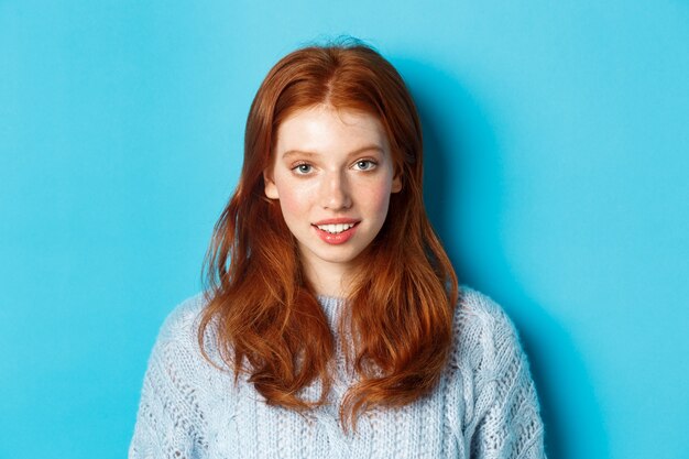 Gros plan d'une jeune fille rousse mignonne souriant à la caméra, debout sur fond bleu.