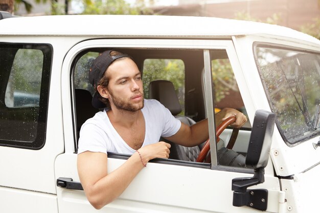 Gros plan d'un jeune homme attrayant avec barbe assis dans son véhicule blanc à la recherche de l'extrême pendant le voyage safari. Homme en snapback conduite sur route rurale