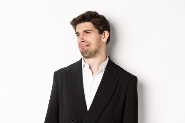 Gros plan sur un jeune homme dégoûté en costume à la mode, grimaçant contrarié, regardant à gauche et debout sur fond blanc.