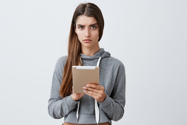 Gros plan de jeune jolie fille brune caucasienne aux cheveux longs en tenue décontractée, lire les nouvelles sur tablette numérique, avec une expression confuse. Concept de mode de vie.