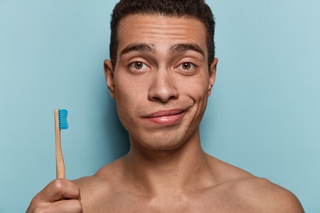 Gros plan d'un jeune avec une peau saine, un corps solide, tient une brosse à dents, va avoir des procédures d'hygiène matinales, se tient contre le mur bleu. Concept d'hygiène, de soins dentaires et de beauté